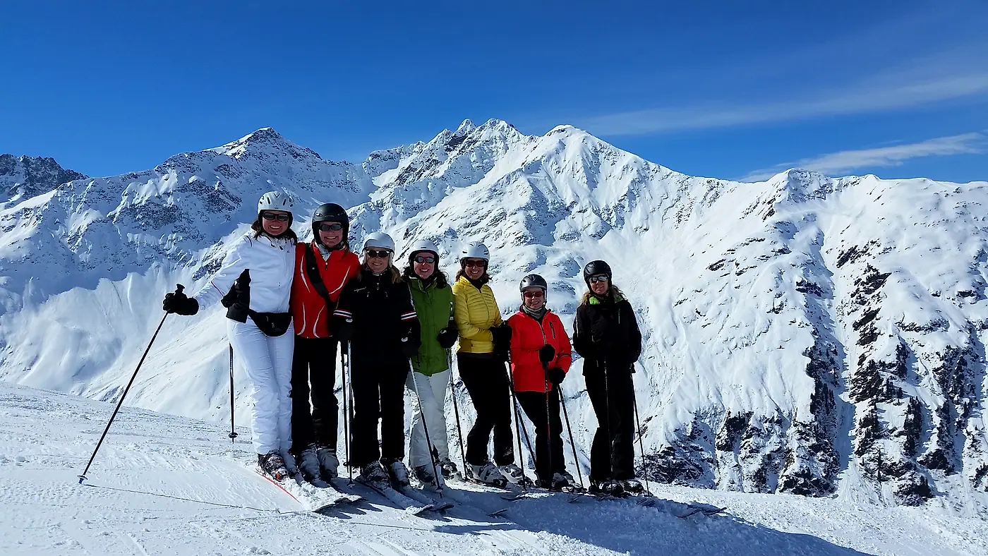 Sieben Skifahrer auf einem Gipfel, dahinter verschneite Berge unter blauem Himmel